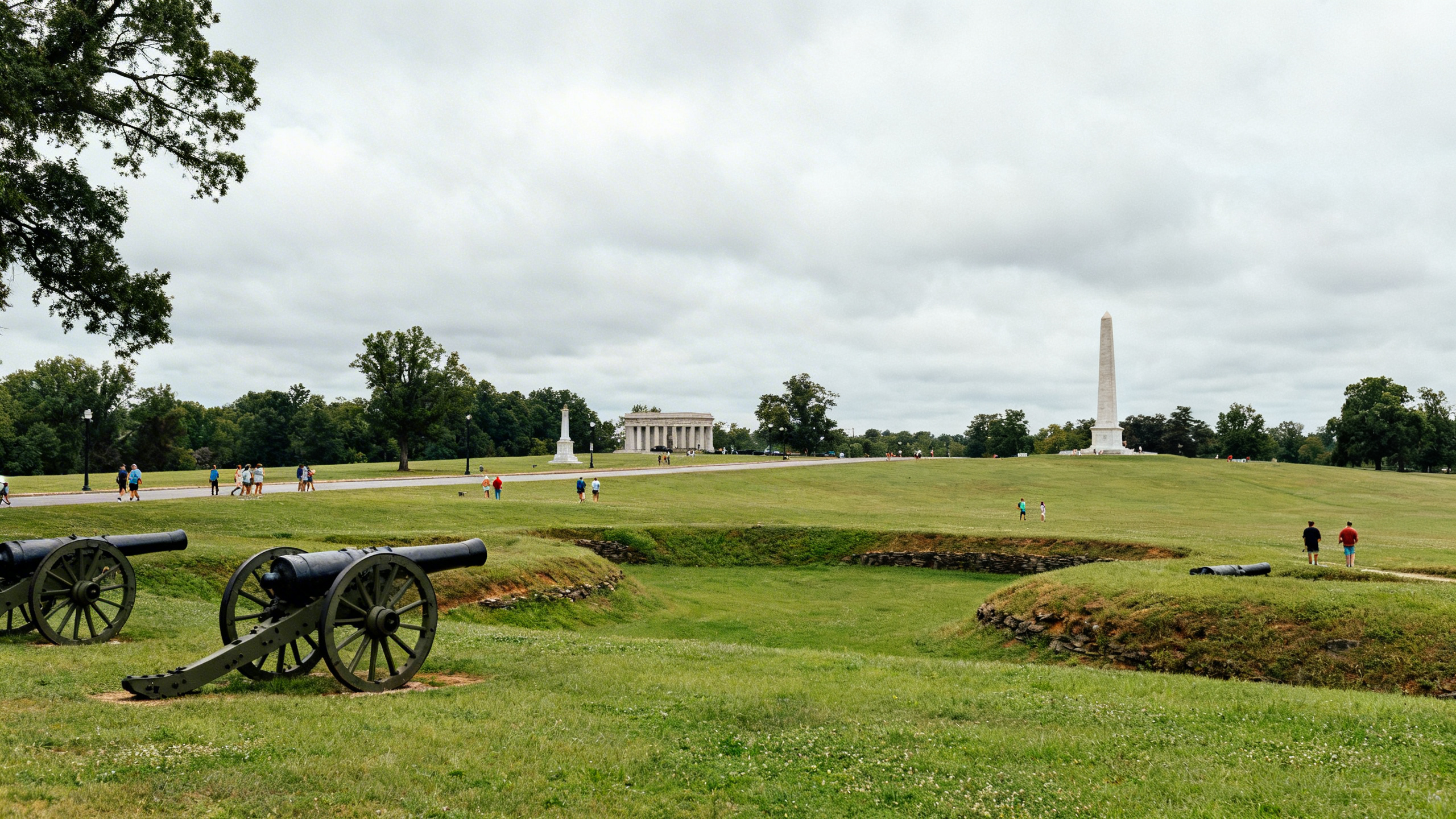 Expansive green fields and preserved Civil War earthworks with large cannons and monuments in the background on a cloudy day