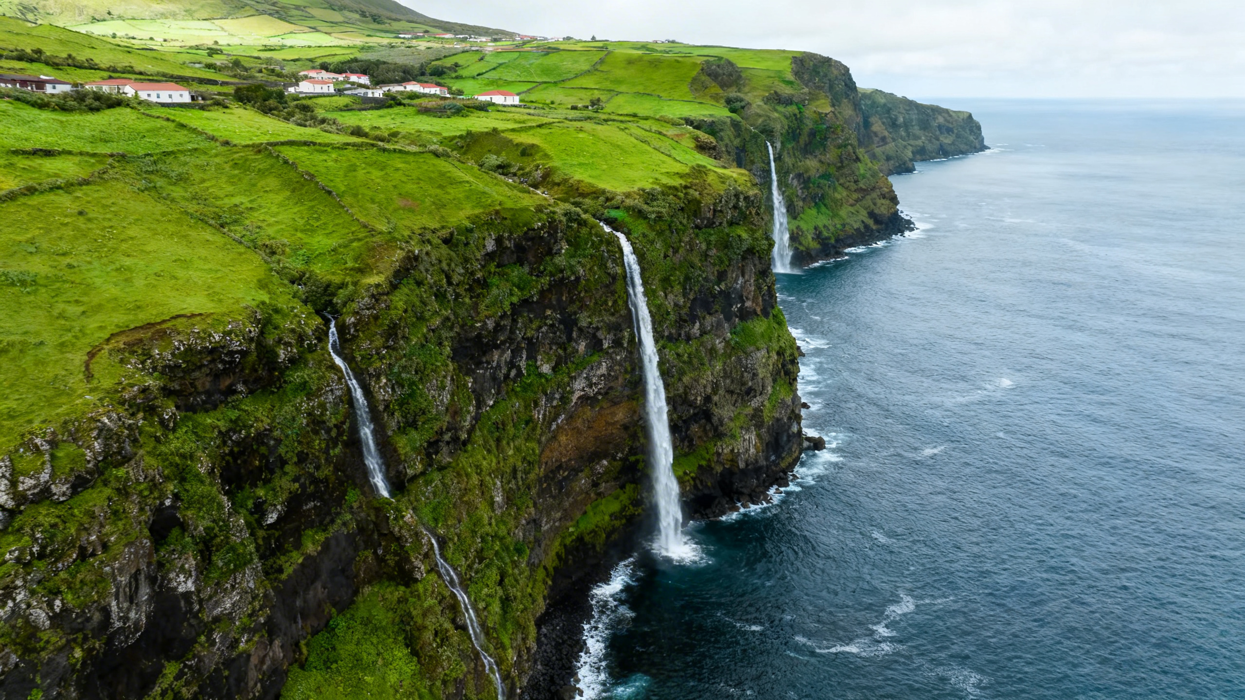 Vivid green cliffs and waterfalls drop sharply into Atlantic Ocean near remote village on Flores island in the Azores archipelago