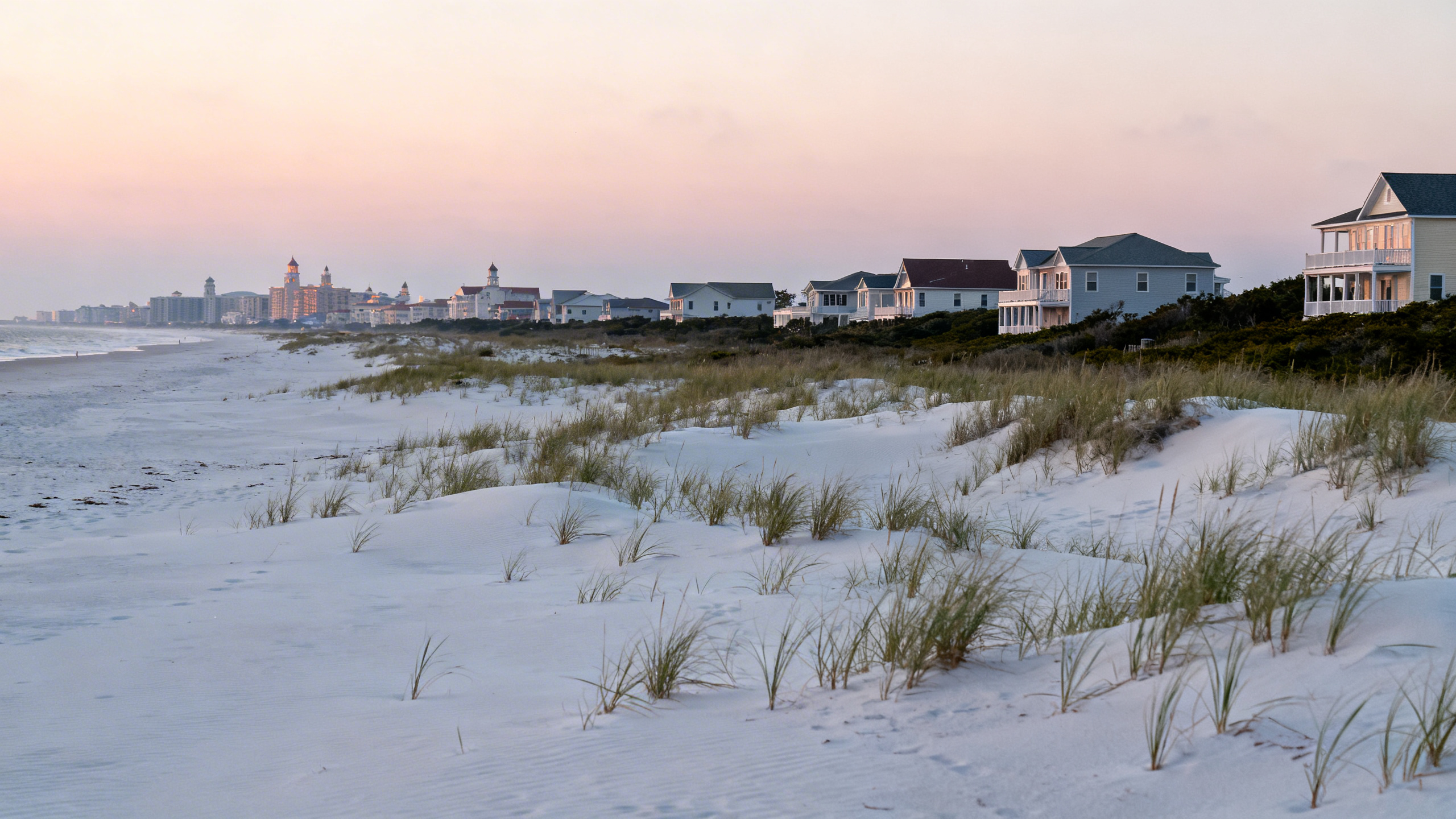 Sandy dunes with sparse coastal grasses and scattered beach houses under a pastel sunset sky in Outer Banks.