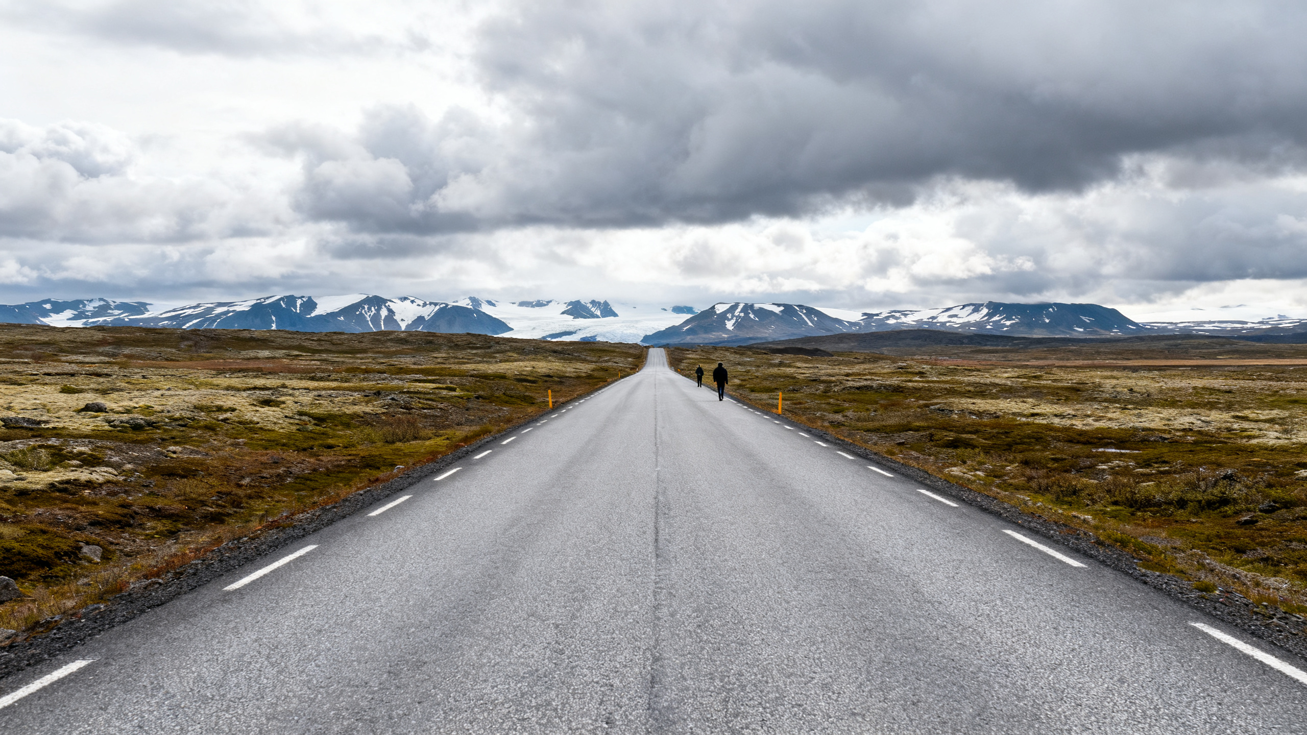 A grey paved highway cuts through a tundra landscape under heavy clouds, with a distant glimpse of snow-capped mountains.