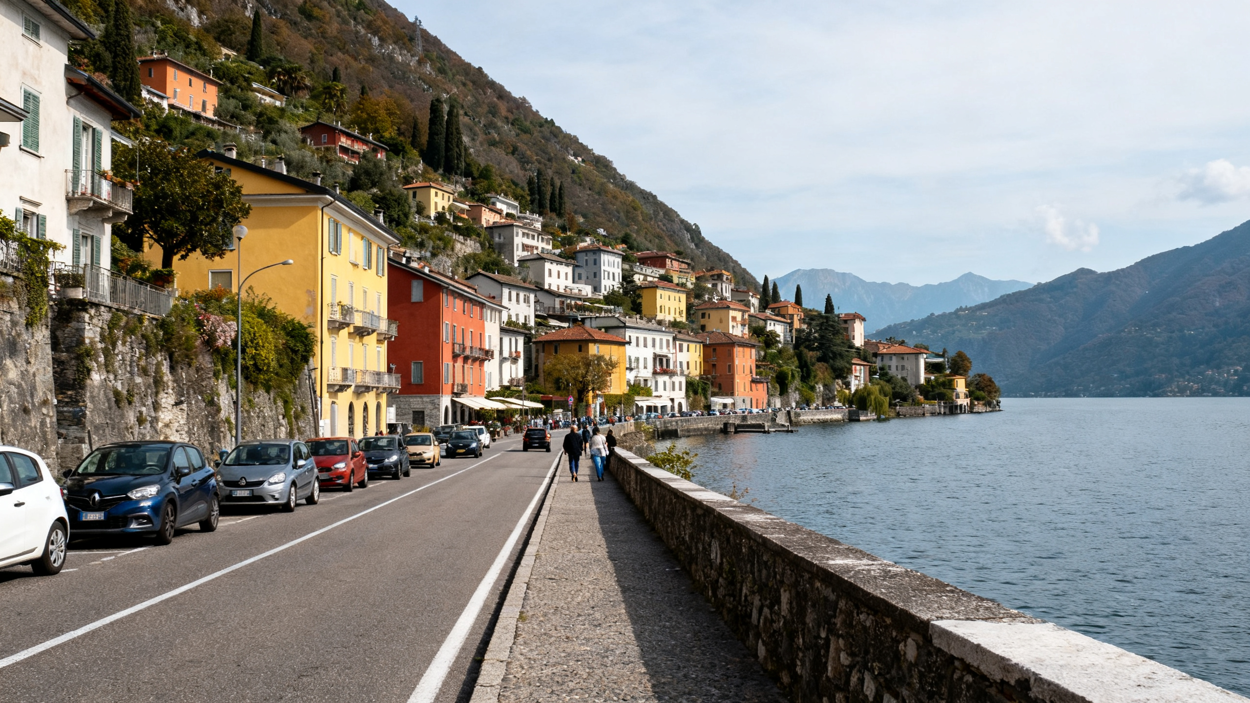 A narrow lakeside road clings to steep hills dotted with colorful buildings and crowded with passing cars during daylight.