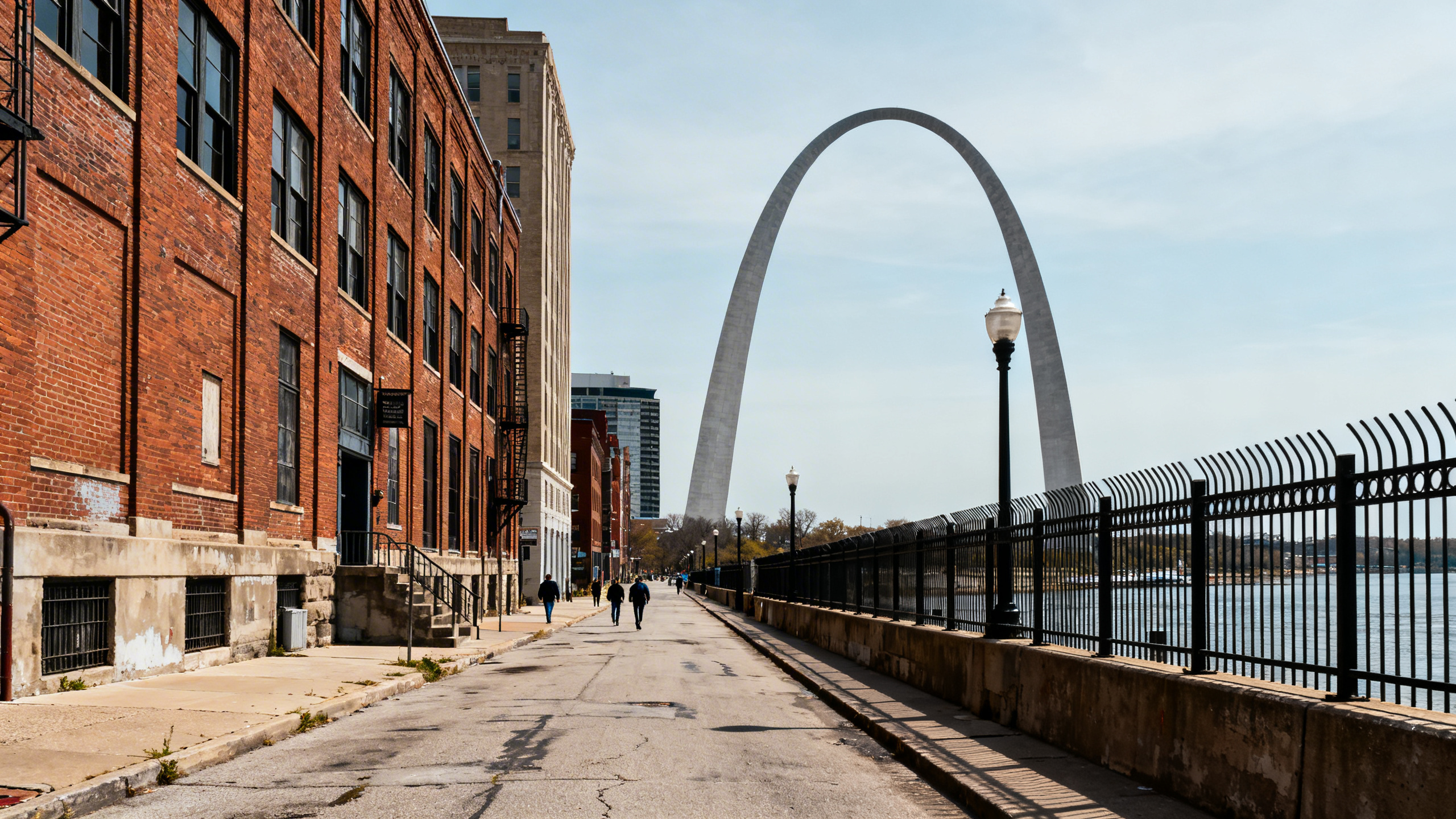 The iconic Gateway Arch towering over a gritty urban riverside street with brick buildings and iron fences