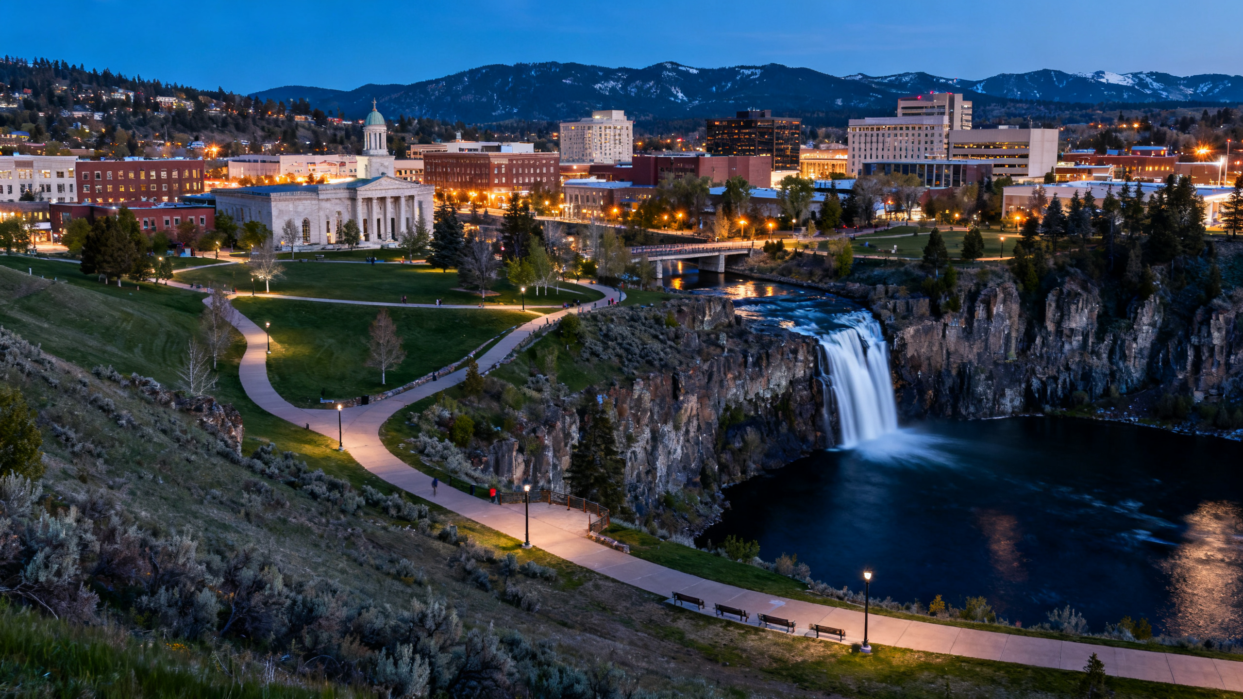 A broad city park with waterfalls and walking paths surrounded by mixed urban and mountain views in Spokane