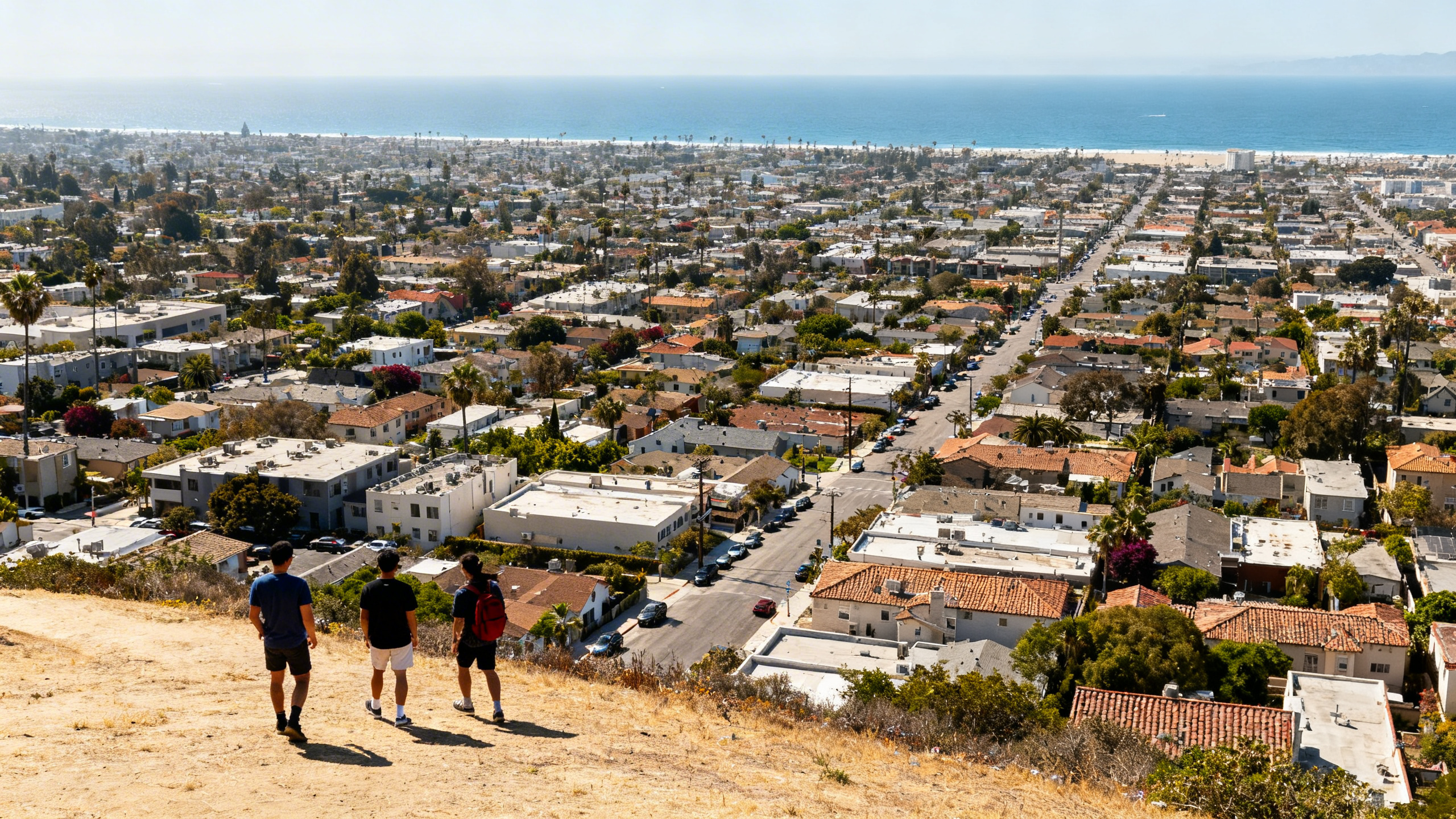 Wide aerial shot of sprawling Los Angeles neighborhoods with the Pacific Ocean visible in the distance on a sunny day