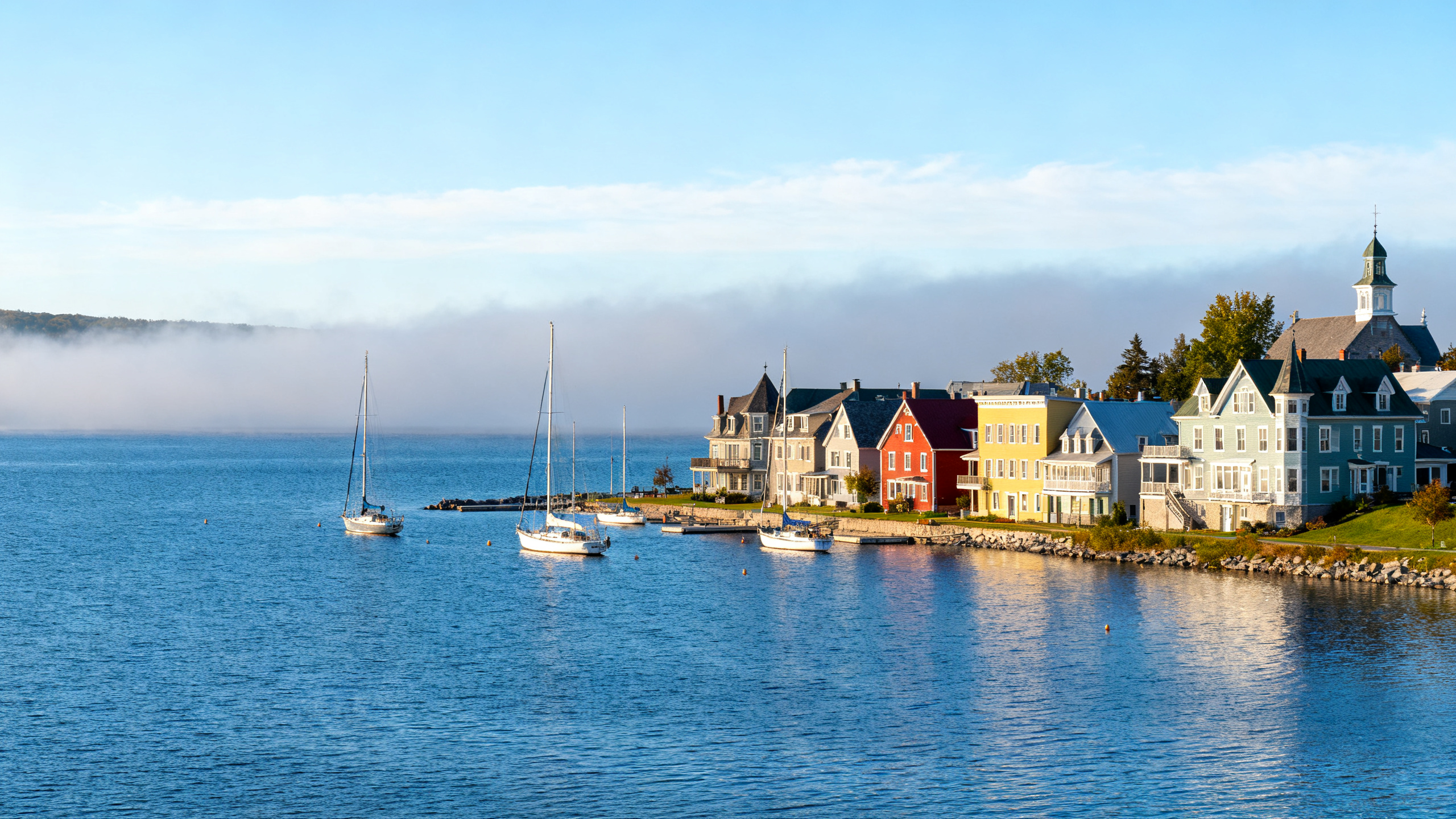 A large blue lake bordered by colorful homes and sailboats under a bright sky in the small town of Charlevoix.