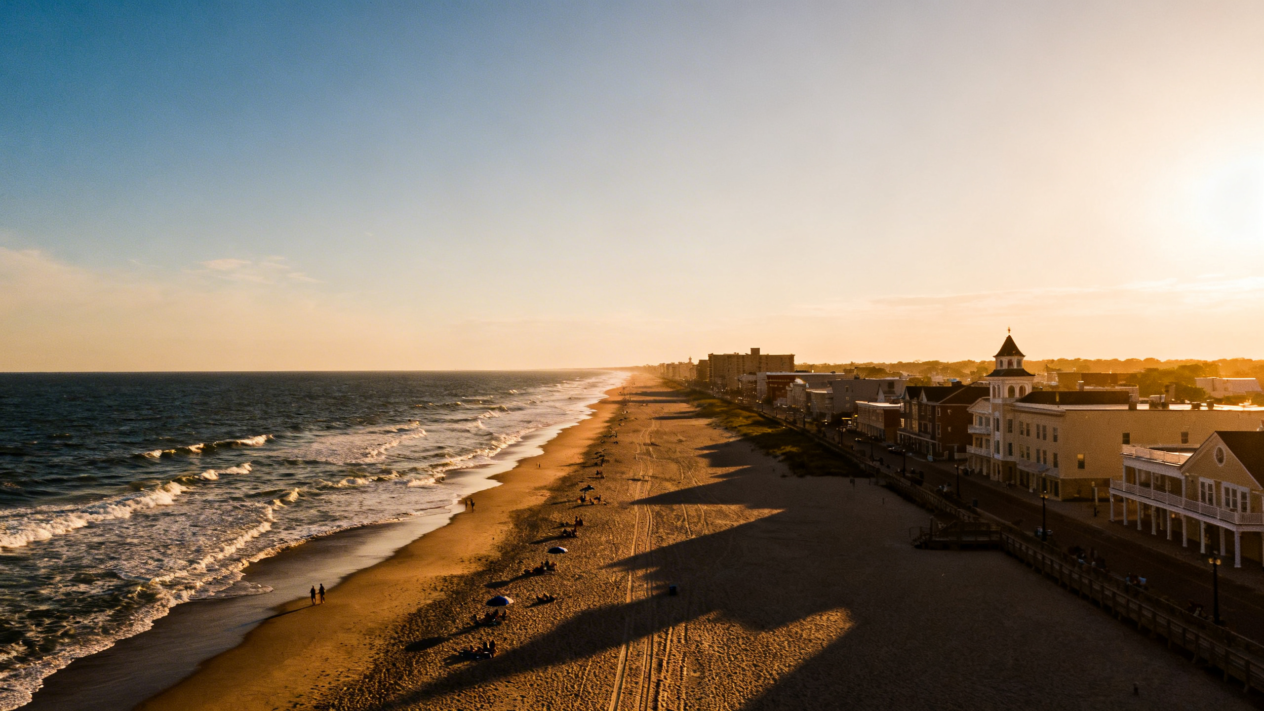 Long stretch of beach with sunbathers and waves, bordered by a low-rise cityscape in Virginia Beach.