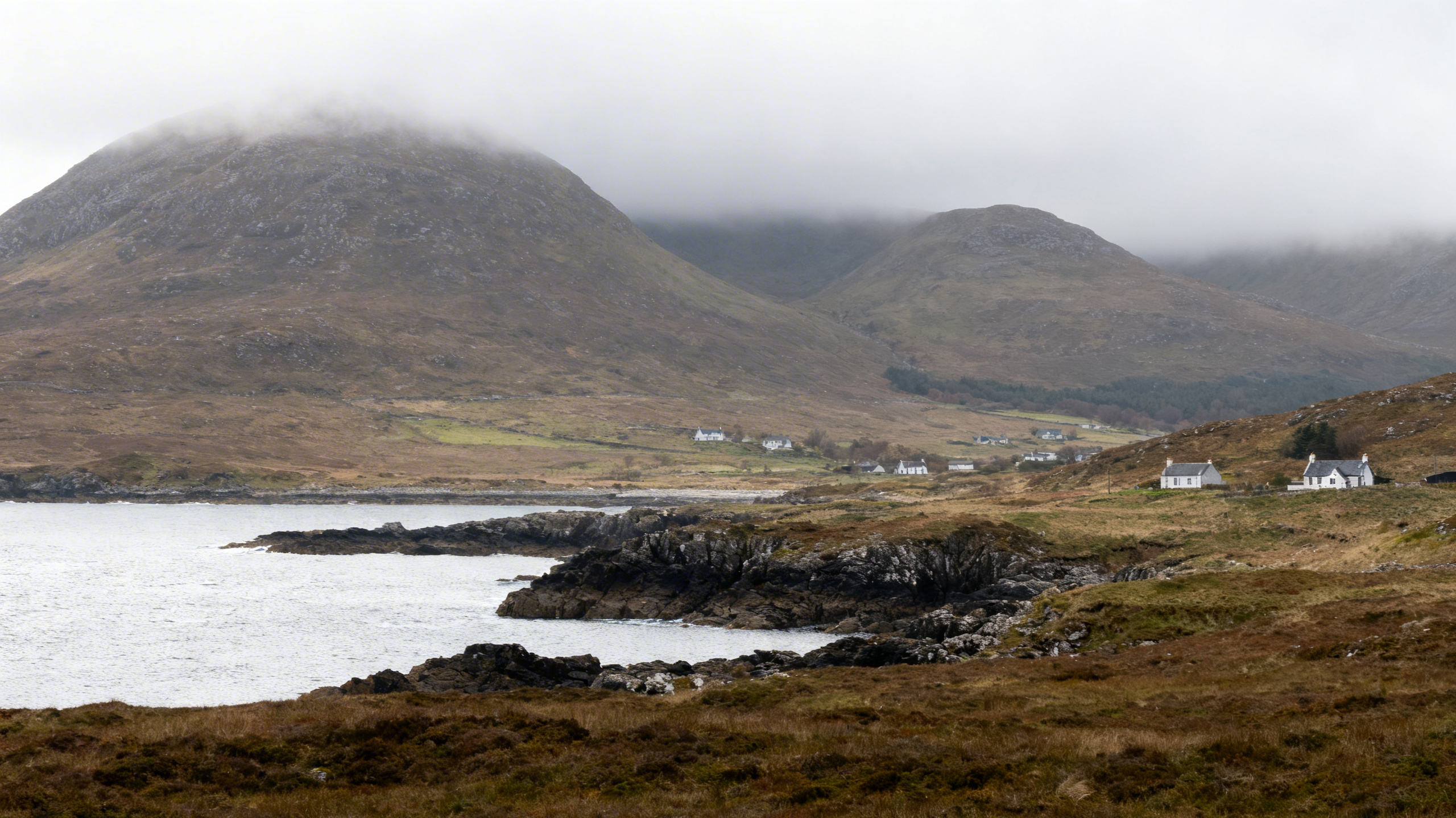 Mist-covered hills rise behind rugged coastline and sparse settlements on the Scottish Isle of Jura, wild and windswept environment
