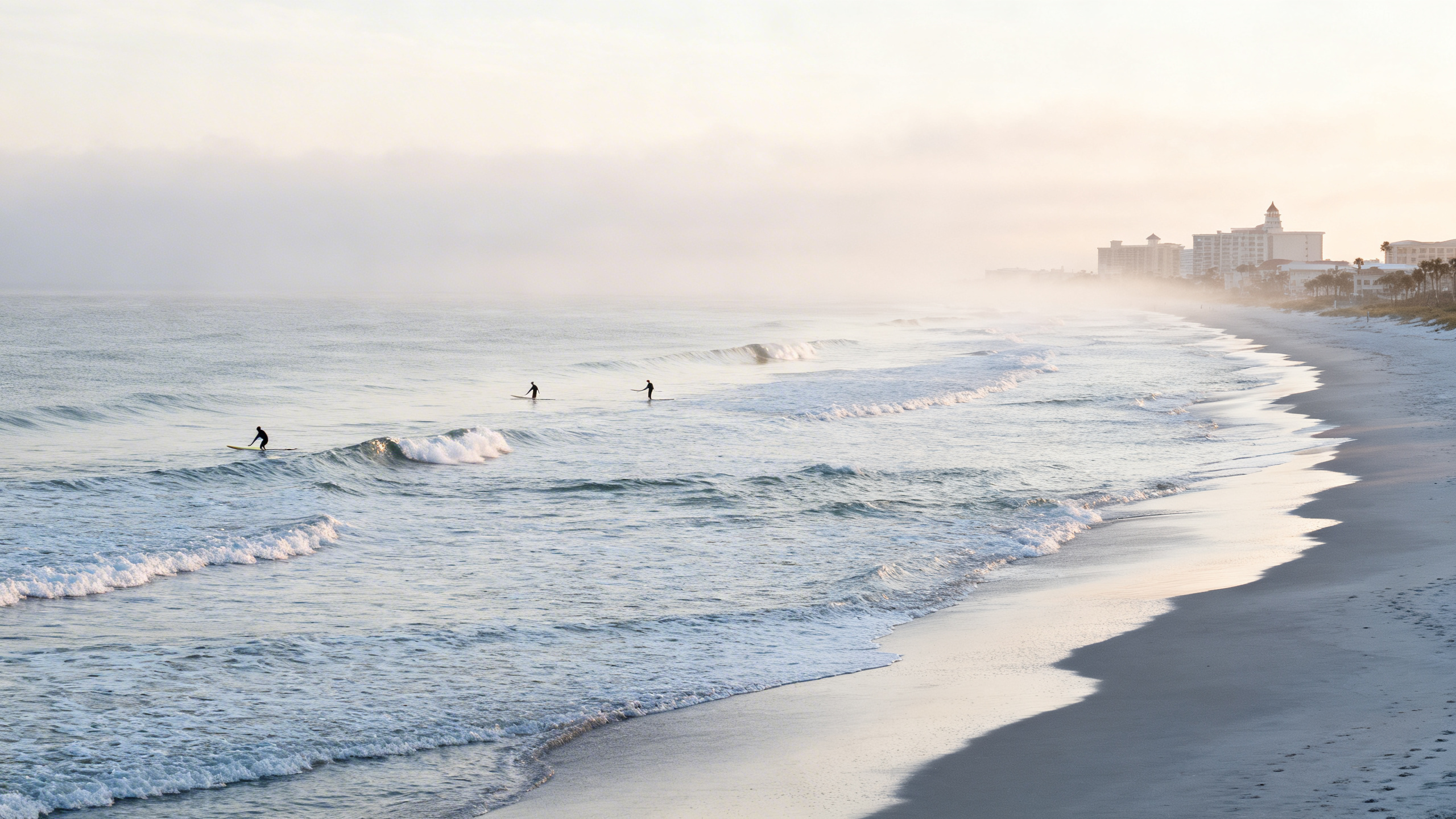 Wide, quiet beach with gentle waves and surfers in the water along New Smyrna Beach shoreline.